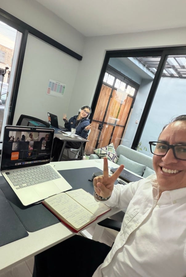 Person in white shirt making peace sign at desk with laptop, another person working at computer in background office space