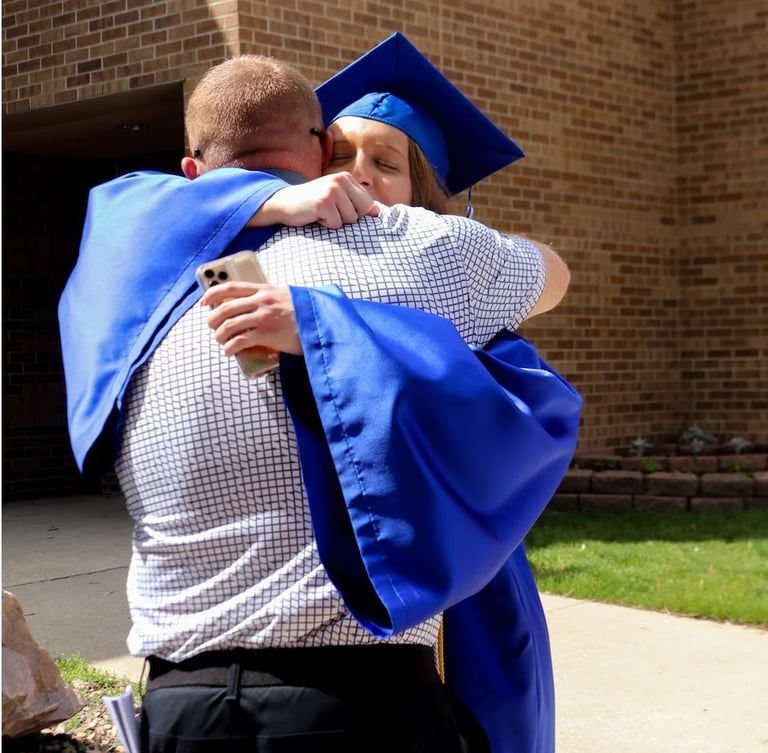 Graduate in blue cap and gown hugging an adult in checkered shirt outside a brick building