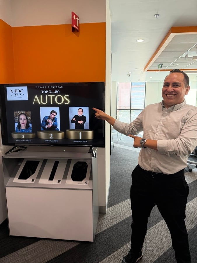 Man in beige shirt pointing at a TV screen displaying Top 3-30 Autos content in an office with orange wall