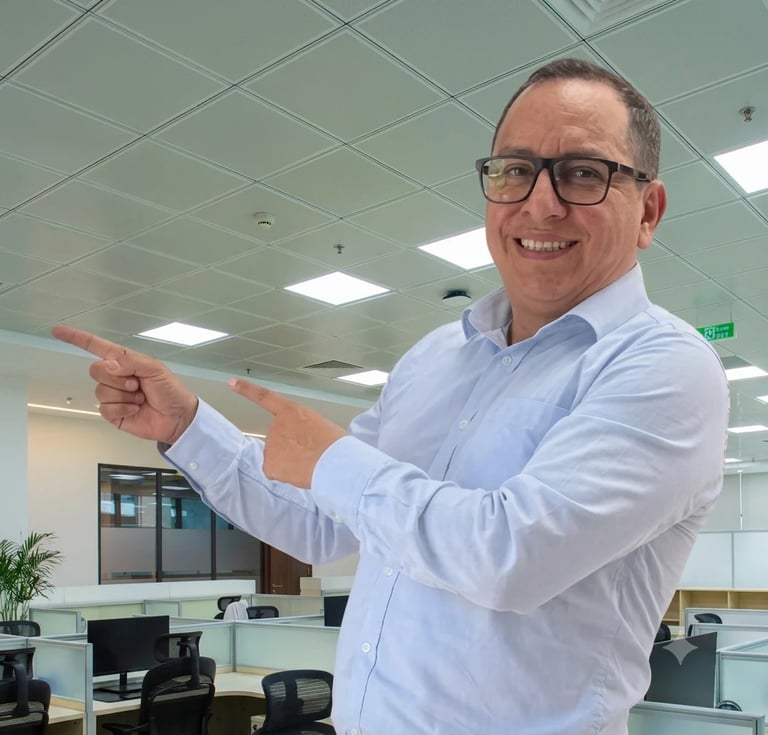 Man in light blue shirt and glasses pointing toward the right, smiling in a modern office environment with suspended ceiling and workstations